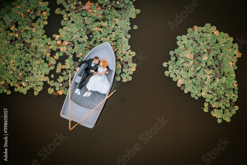 Aerial view of a couple in wedding attire lying in a small rowboat on a pond surrounded by lily pads, creating a romantic and serene scene. photo