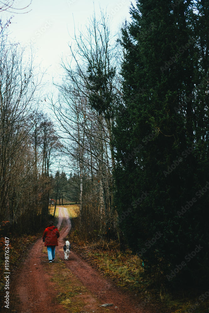 Woman in a red jacket walking a dog on a forested dirt path in w