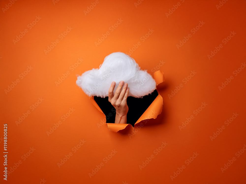 Fototapeta premium A hand emerges from a torn orange background, holding a fluffy white cloud. The scene is surreal and playful, suggesting imagination or a breakthrough.