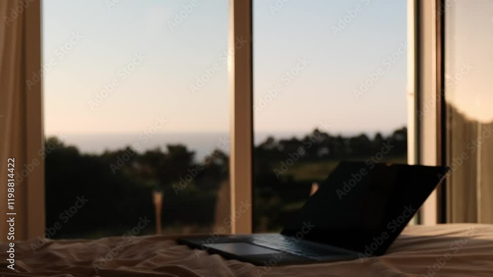 Laptop on bed with open window overlooking scenic greenery and ocean Laptop on bed with open window overlooking scenic greenery and ocean