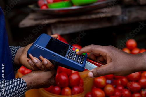 A hand using a bank card on a point-of-sale terminal at a market stall selling fresh tomatoes, representing digital financial transactions and modern commerce in traditional outdoor settings. photo