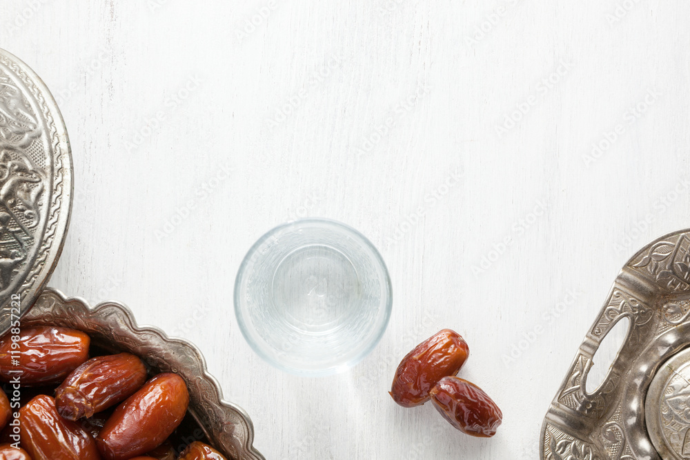 Top view on silver plate with date fruits and glass of water on a white wooden background. Ramadan background.