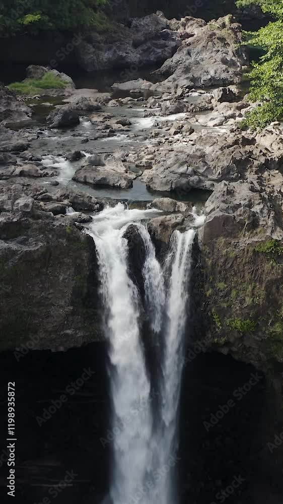 Aerial view of Rainbow Falls on the Big Island, Hawaii, surrounded by lush green foliage and rugged rocky terrain. Aerial view of Rainbow Falls on the Big Island, Hawaii, surrounded by lush green foliage and rugged rocky terrain.