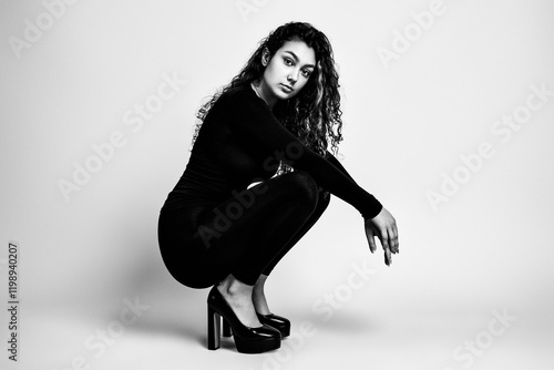 Black and white portrait of a beautiful teenage girl in a tight black suit and black high-heeled shoes against a homogeneous background photo