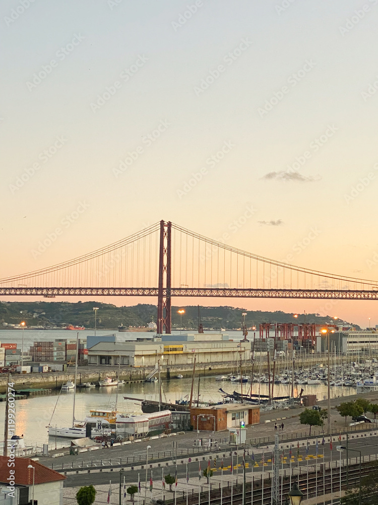 Fototapeta premium Sunset view of the iconic red suspension bridge over the river, marina with yachts, industrial port, and clear sky.