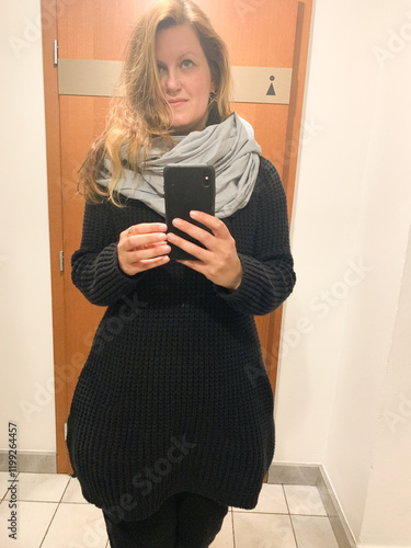 Woman capturing a mirror selfie in a cozy black sweater and light scarf,