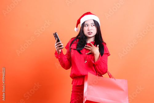 beautiful girl charm wearing christmas dress, santa hat, afraid to look left, hands dumbfounded using gadget, carrying red bag of christmas gifts, isolated on orange background photo