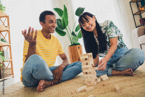 Photo of lovely cute charming positive multinational couple spend weekend sit floor play jenga build tower pile stack indoors photo