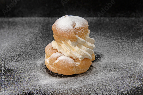 A cream puff from a bakery, photographed against a black background. The cream puff is dusted with powdered sugar, highlighting its texture and the generous amount of whipped cream filling photo