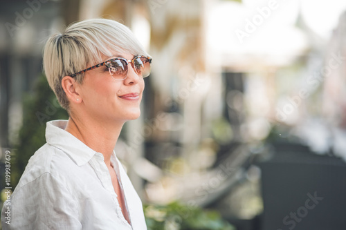 Gorgeous woman with blond short hair looking to side. Face of female person looking to side dressed in white summer shirt. Outdoors, blurred street in the city.