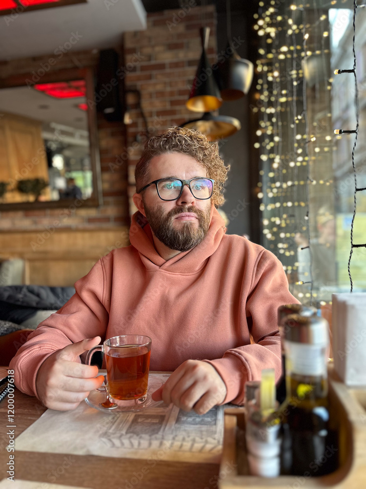 Bearded man in a peach hoodie enjoys a glass of tea in a cozy cafe. Warm string lights and rustic decor, creating a relaxed and intimate mood in restaurant Bearded man in a peach hoodie enjoys a glass of tea in a cozy cafe. Warm string lights and rustic decor, creating a relaxed and intimate mood in restaurant