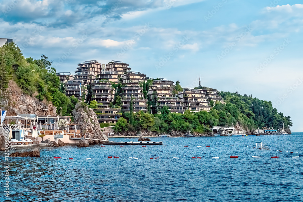 Naklejka premium A scenic coastal view of Dukley Gardens in Budva, Montenegro, with luxury residential buildings perched on a hillside overlooking the Adriatic Sea. The waterside resort area with a restaurant