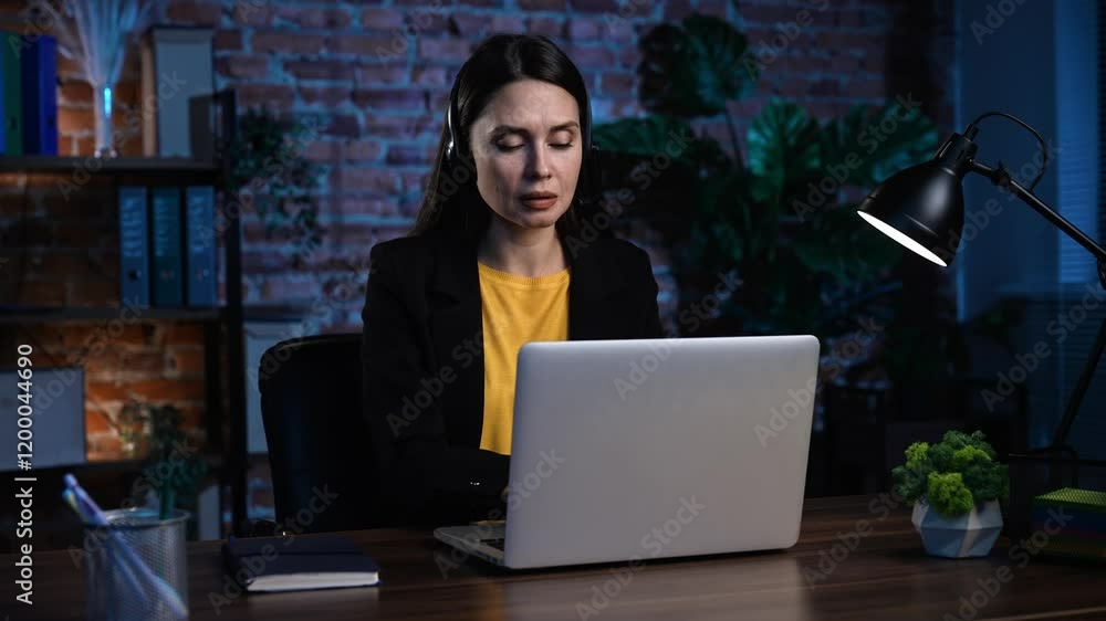 Young business woman sitting at desk working late in office loft, using laptop, wearing headset talking with client, typing on keyboard, checking documentation. Young business woman sitting at desk working late in office loft, using laptop, wearing headset talking with client, typing on keyboard, checking documentation.