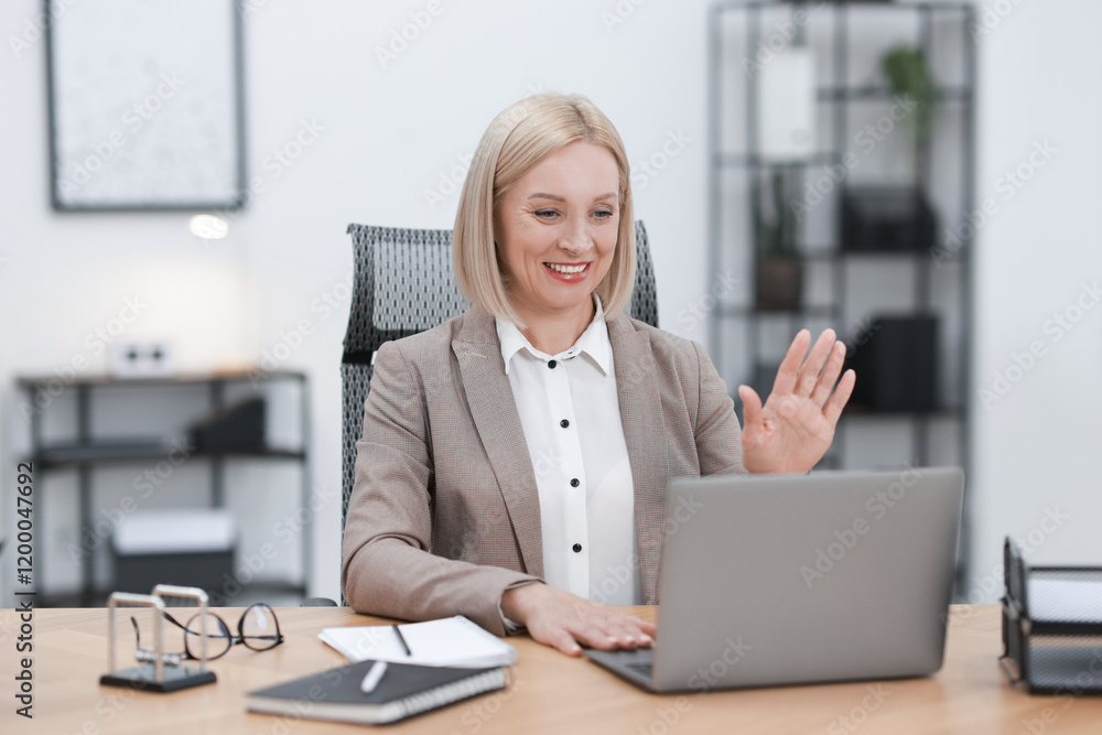 Smiling middle aged woman having videochat by laptop at table in office Smiling middle aged woman having videochat by laptop at table in office