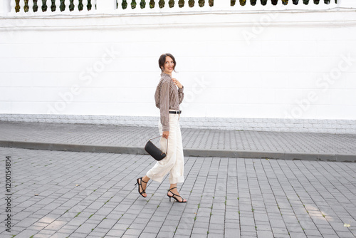 Young beautiful brunette woman wearing silk striped brown shirt, white denim pants, black heeled sandals and small handbag walking outdoor on a city street. Trendy casual outfit. Street fashion.