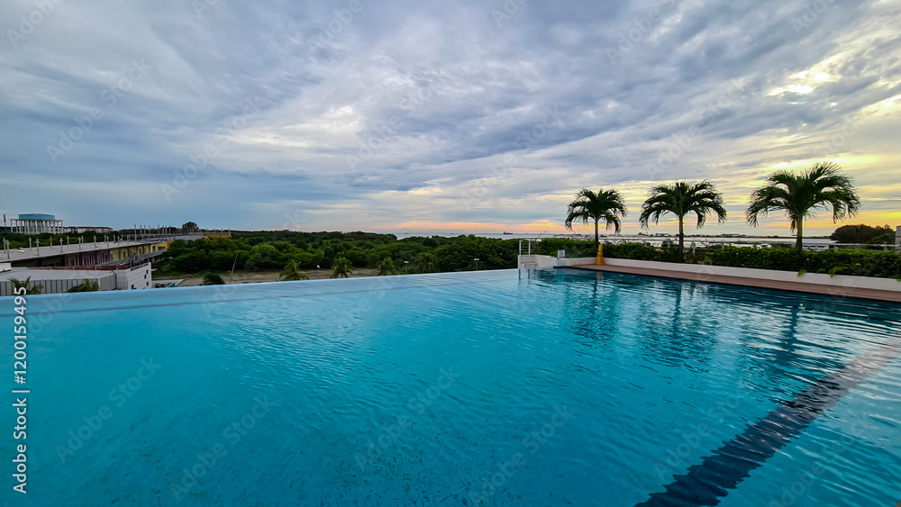 Naklejka premium Infinity pool with calm, blue water reflects cloudy sky. Palm trees near pool's edge, overlooking coastal landscape with greenery and buildings in distance. Tranquility and luxurious vacation setting