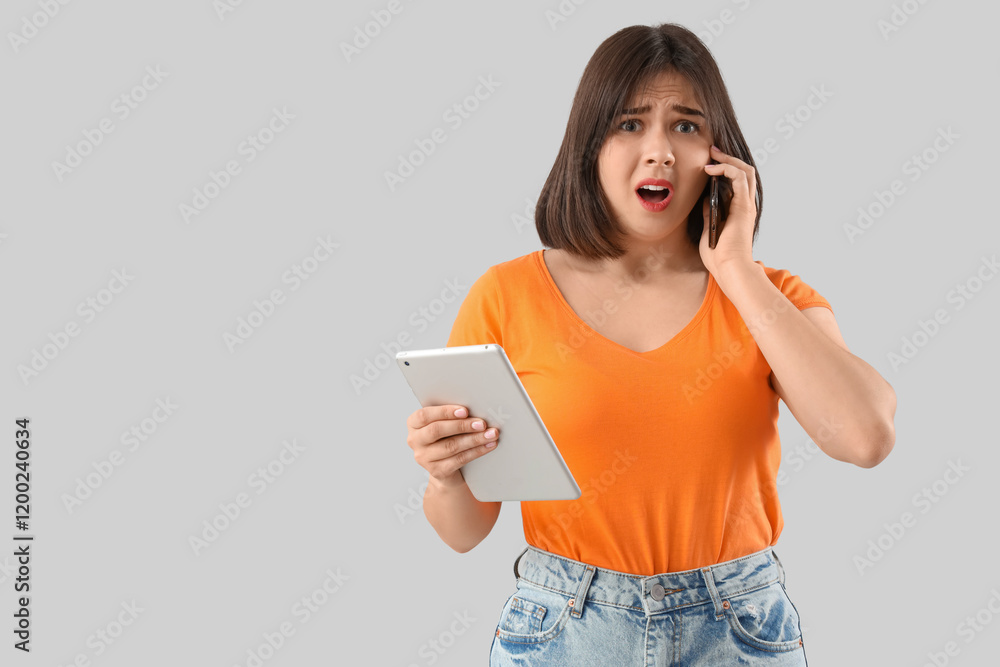 Young stressed woman with tablet talking by mobile phone on grey background. Stress Awareness Month Young stressed woman with tablet talking by mobile phone on grey background. Stress Awareness Month