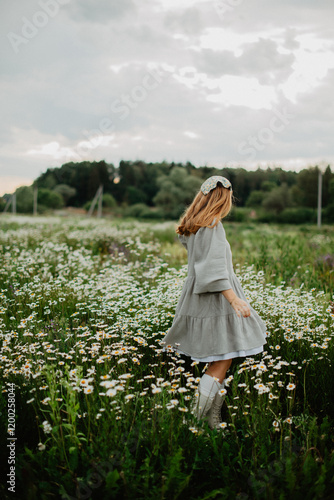 A young woman in a flowy dress and headband walks through a blooming daisy field, enjoying the peaceful countryside atmosphere under a cloudy sky.
