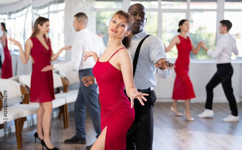 Fototapeta premium Cheerful stylish duet, expressive African American man in formal wear and elegant woman in red dress dancing vintage twist in modern dance studio. Active hobbies concept..