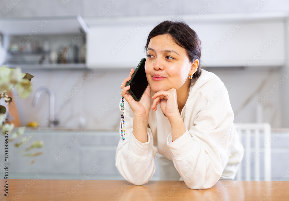Positive relaxed young dark-haired woman dressed in casual white clothing standing leaning on table in home kitchen, having friendly conversation on smartphone.. Positive relaxed young dark-haired woman dressed in casual white clothing standing leaning on table in home kitchen, having friendly conversation on smartphone..