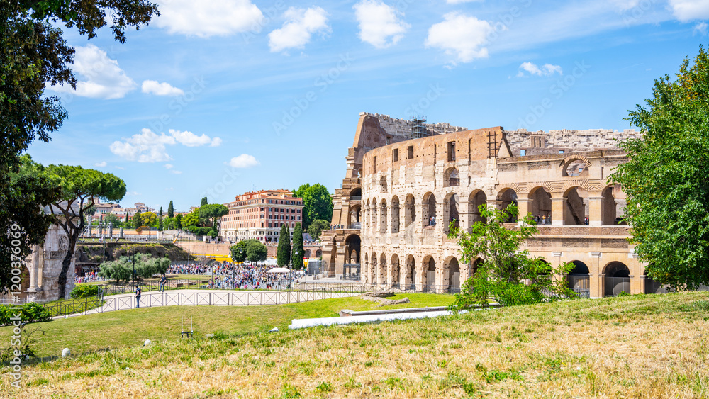 Fototapeta premium Visitors enjoy the historic Colosseum in Rome, Italy on a bright day. The ancient structure stands majestically against a backdrop of lush greenery and blue skies, showcasing its timeless beauty.