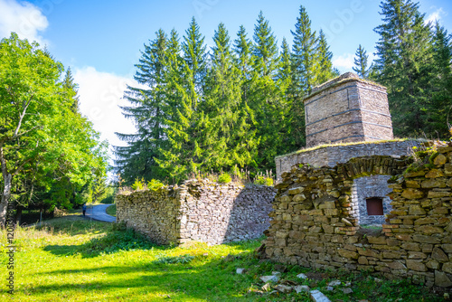 The historic remains of a lime kiln stand surrounded by lush greenery in the Ore Mountains near Kovarska. Sunlight filters through towering trees, highlighting the stone structure. photo