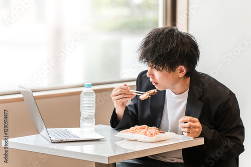 イートインスペース・社員食堂でパソコンを見ながら昼食にお弁当を食べる若いアジア人男性ビジネスマン
 photo