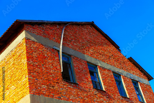 Red brick building with blue-framed windows under a clear blue sky. The structure shows rustic textures, sunlight highlights, and a simple architectural style, evoking an urban, vintage feel