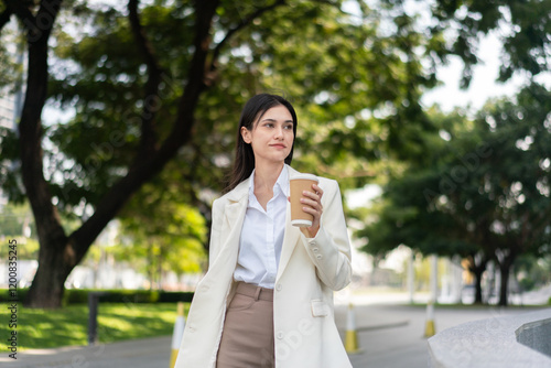 Confident woman in formal attire walking outdoors with a coffee cup in hand, enjoying a stroll through a green park setting.