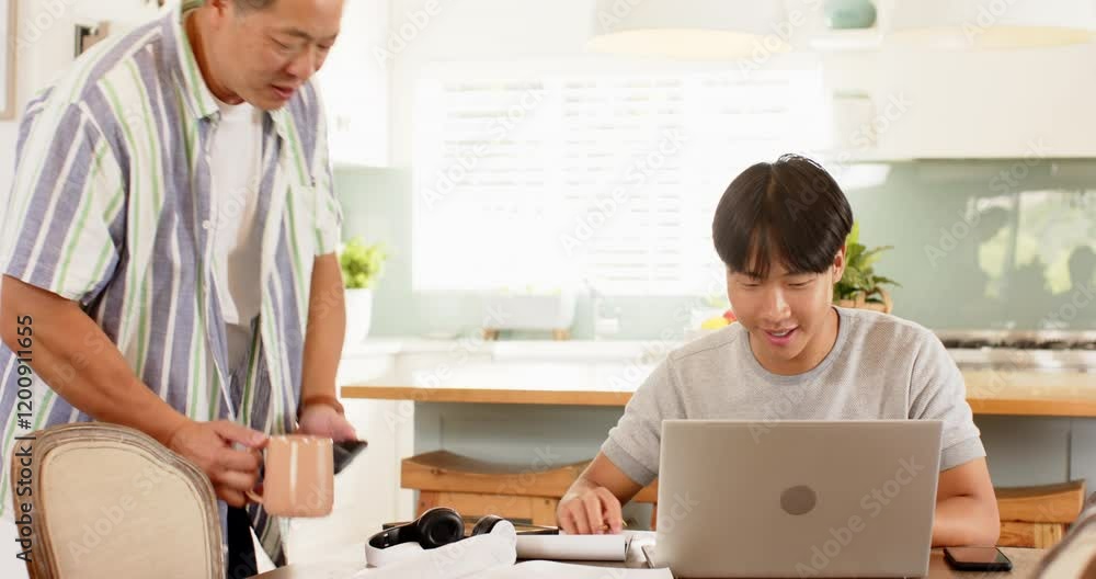 Working from home, young Asian man using laptop while older Asian man talking to him Working from home, young Asian man using laptop while older Asian man talking to him