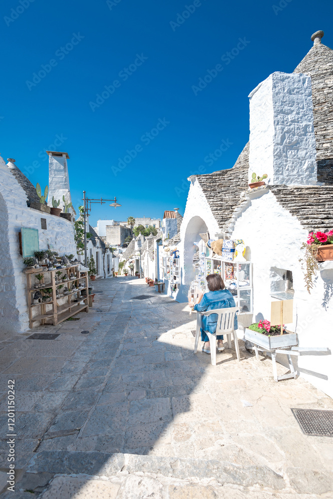 Beautiful stone of Trulli houses with white walls, narrow streets, decorated with flowers, palm trees and other decorations. Spring day, warm with sun and blue skies Selling souvenirs