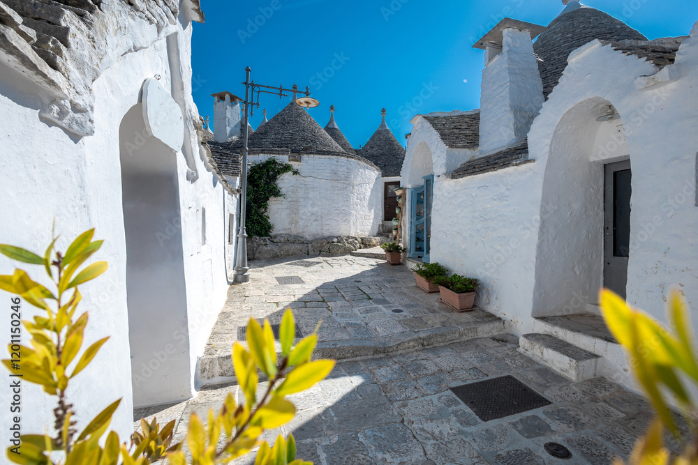 Beautiful alley of Trulli houses with white walls, narrow streets, decorated with flowers, palm trees and other decorations. Spring day, warm with sun and blue skies Selling souvenirs
