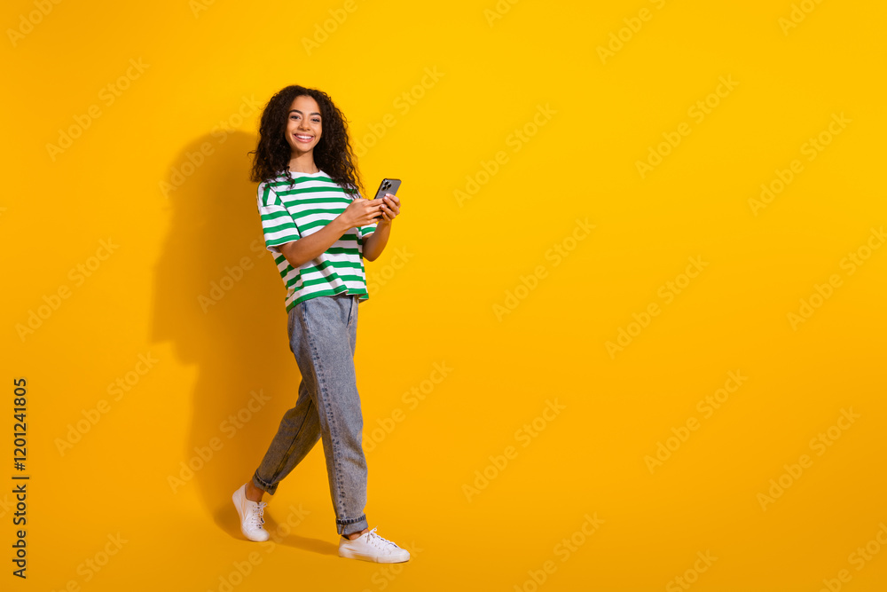 Full size photo of pretty young girl walk hold phone wear striped t-shirt isolated on yellow color background Full size photo of pretty young girl walk hold phone wear striped t-shirt isolated on yellow color background