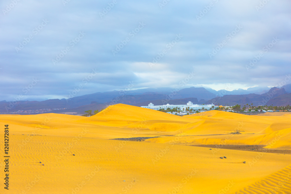 Golden sand dunes of Maspalomas, Canary Islands, stretching towards horizon with a resort in distance. Mountains and cloudy skies frame tranquil desert landscape. Perfect for travel and nature themes