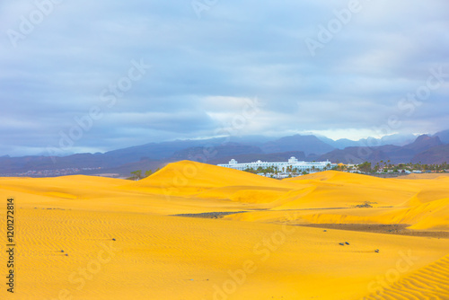 Obraz na płótnie Golden sand dunes of Maspalomas, Canary Islands, stretching towards horizon with a resort in distance. Mountains and cloudy skies frame tranquil desert landscape. Perfect for travel and nature themes w salonie