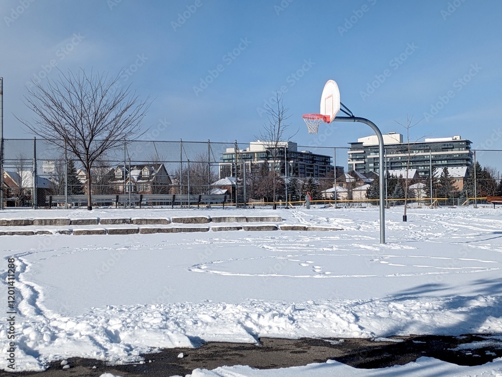 Fototapeta premium Snow covered basketball court at River Glen Park in Oakville, with baseball field , trees and apartment buildings in the background, during a cold winter day with blue sky