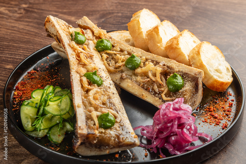 Fotografie A delicious serving of bone marrow topped with herbs, accompanied by toasted bread, pickled onions, and sliced cucumbers, presented on a black plate against a wooden background
