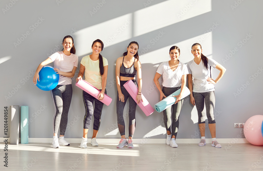 Fototapeta premium A group of athletic girls with yoga mats stand against the wall in the gym, portrait. Happy sportswomen smiling. Joy and satisfaction from yoga. Concept of health and perseverance in sports