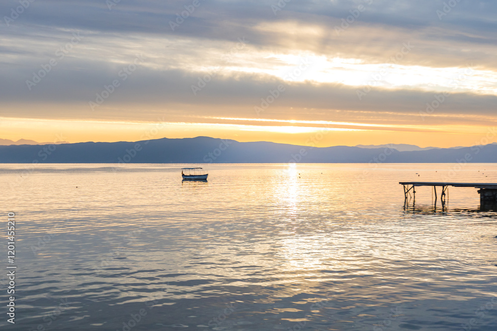 Fototapeta premium Breathtaking sunset over Lake Ohrid, with pier and a small boat anchored in the calm waters. The golden light reflecting on the water creates a serene and picturesque scene.