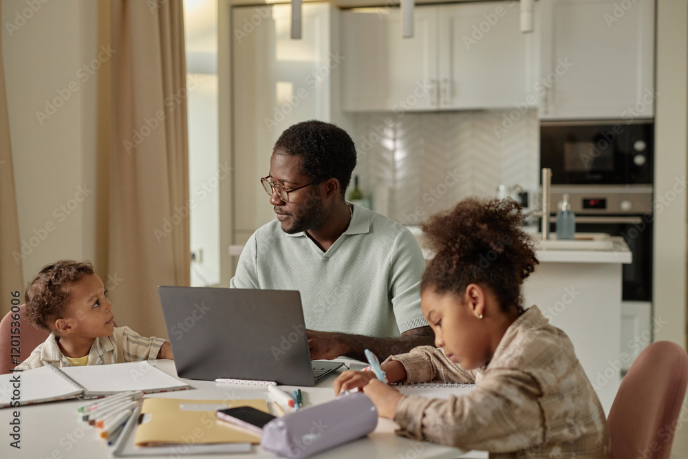 Medium shot of adult African American man talking to little curious son while working on laptop at table with children playing nearby in cozy kitchen, concept of work life balance Medium shot of adult African American man talking to little curious son while working on laptop at table with children playing nearby in cozy kitchen, concept of work life balance