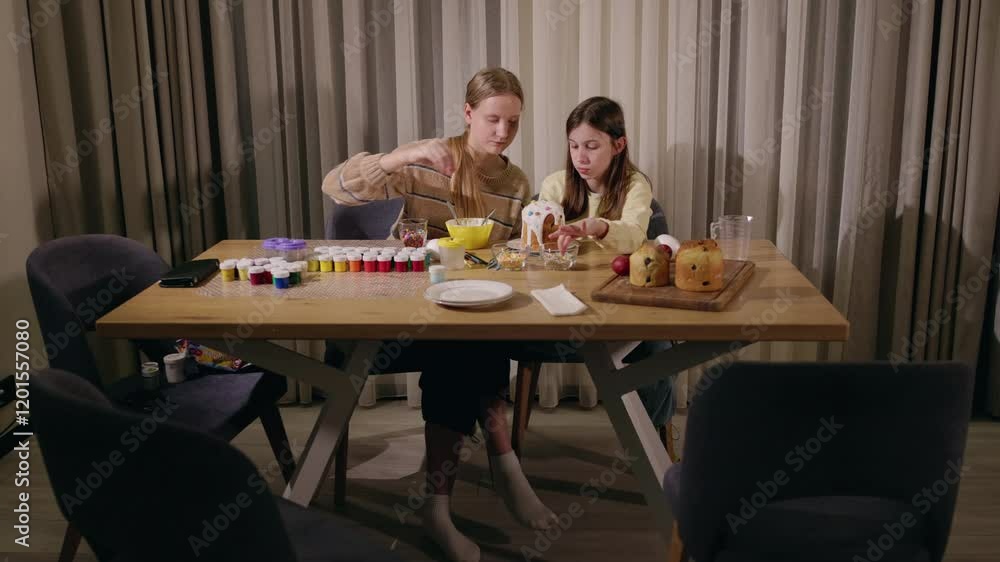 Two girls decorating an Easter cake at a wooden table, surrounded by colorful paints, brushes, sprinkles, and festive supplies, creating a holiday atmosphere. Two girls decorating an Easter cake at a wooden table, surrounded by colorful paints, brushes, sprinkles, and festive supplies, creating a holiday atmosphere.