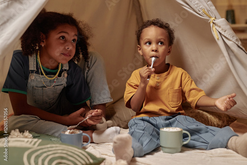 Two African American brother and sister of preschool age sitting inside teepee in bedroom enjoying hot chocolate drinks with marshmallows on top, while looking at parent outside toy wigwam photo