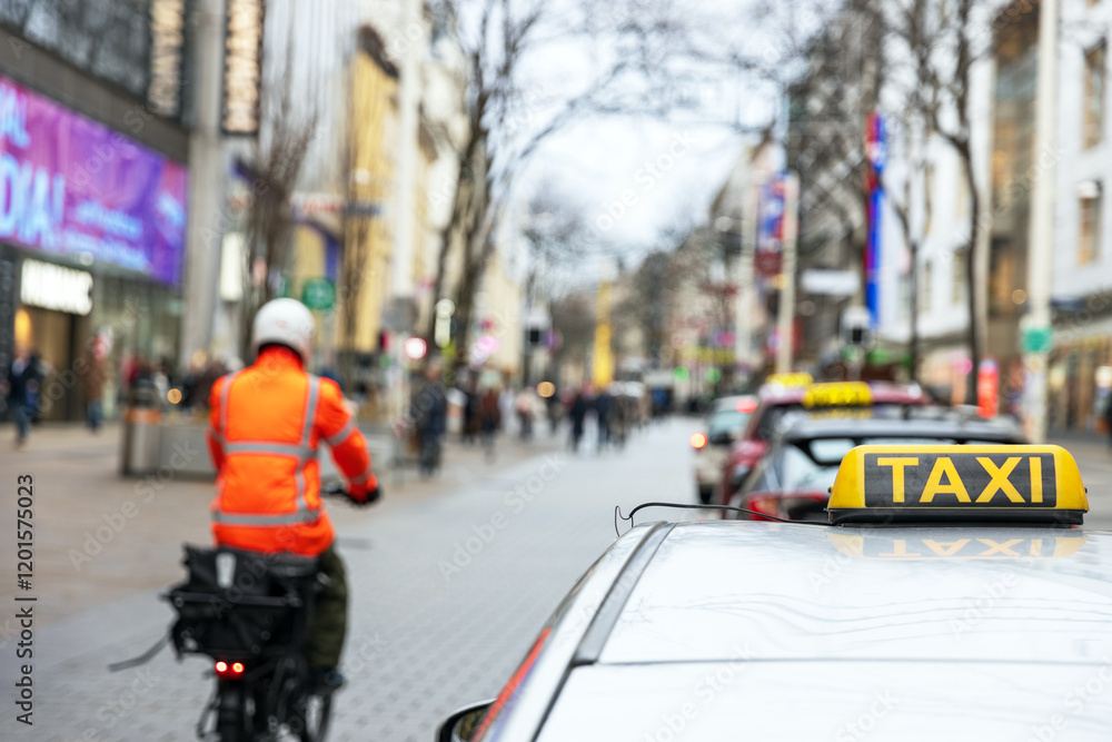 Busy city street scene with a cyclist in orange safety gear and a taxi sign in focus Busy city street scene with a cyclist in orange safety gear and a taxi sign in focus