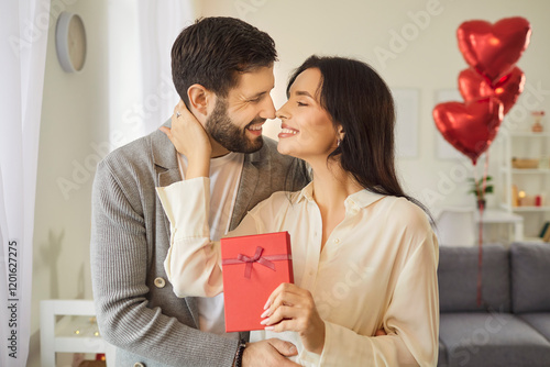 Woman beams with joy as she holds red gift in her hands, celebrating Valentines Day with her man. Loving couple gazes at each other tenderly in living room decorated with heart-shaped balloons. photo