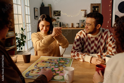 Group of friends looking at young adult woman who smiling while shaking dice, they playing board game at home photo