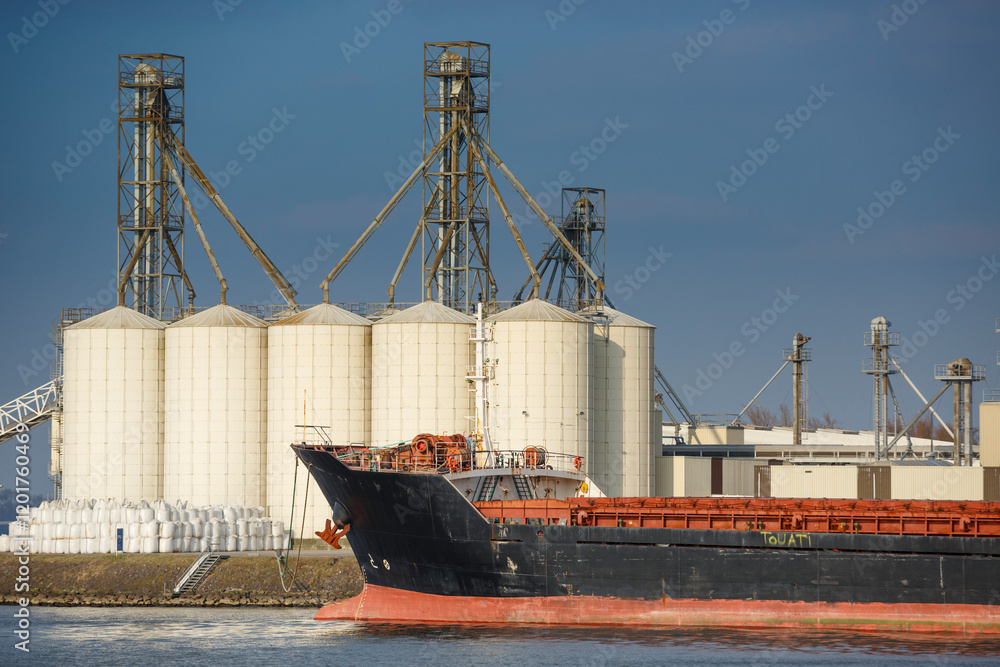 Fototapeta premium Cargo ship named Mahan Tide docked near large grain silos during a clear day with blue skies