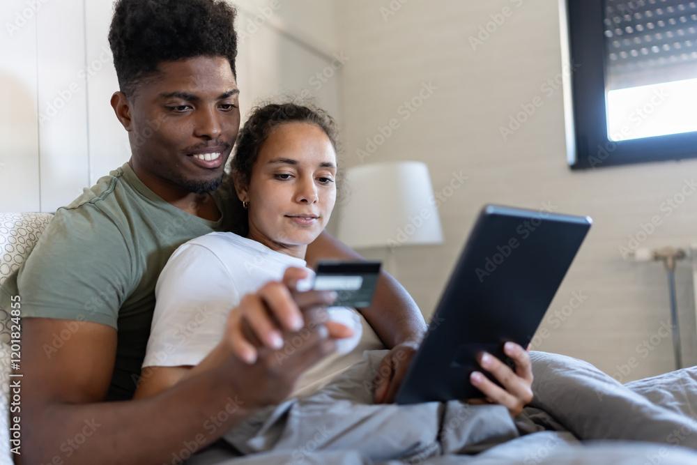 A young couple sits together on a bed, holding a credit card and tablet, smiling as they engage in online shopping in a cozy, well-lit bedroom A young couple sits together on a bed, holding a credit card and tablet, smiling as they engage in online shopping in a cozy, well-lit bedroom