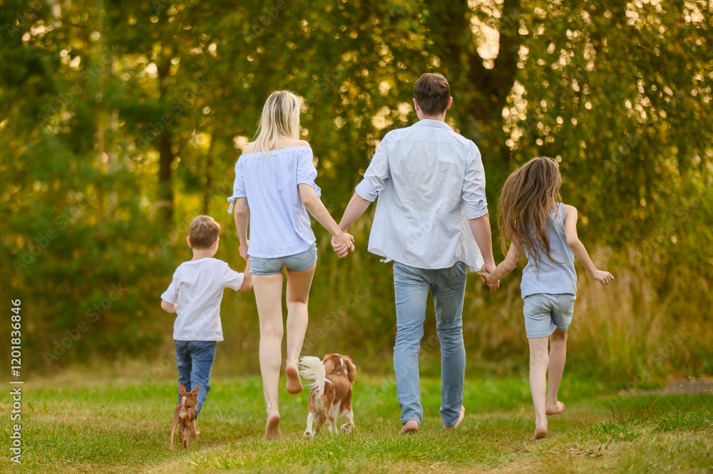 Fototapeta premium Family enjoying beautiful day outdoors, holding hands, running barefoot together on grass, view from back. Family walking happy dogs. Concept of summer weekends, country vacation, being with nature.
