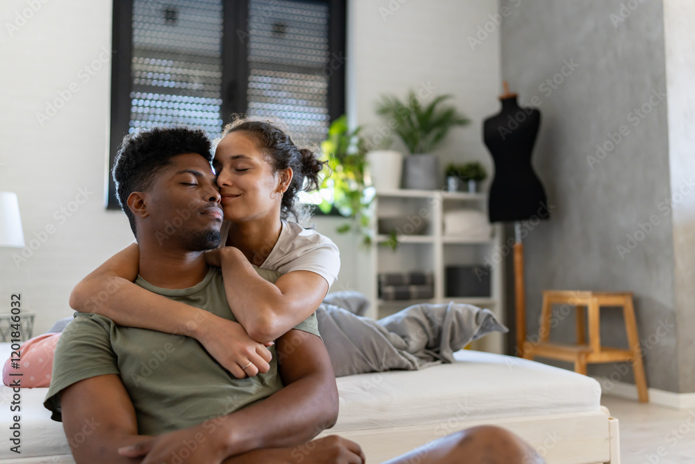 Obraz premium A loving couple sitting on the floor near a bed, the woman embracing the man from behind as he leans back, both sharing an affectionate moment in a cozy bedroom