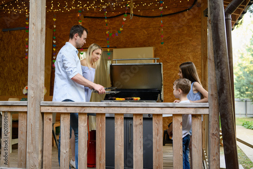 Cheerful family is busy preparing delicious food on grill located on their welcoming veranda, enjoying beautiful atmosphere outdoors. BBQ party in country house.
